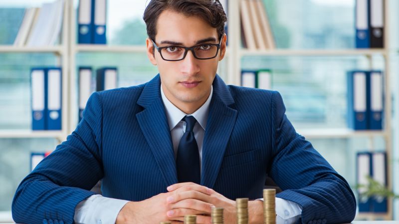 Businessman in a blue suit sitting at a desk with stacks of coins, symbolizing wealth management and salary negotiations.