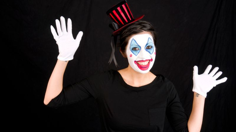 A cheerful person dressed as a clown, wearing a black outfit, white gloves, and a small top hat with red and black stripes.