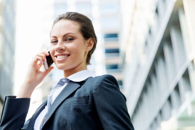 Eine lächelnde Geschäftsfrau im Anzug telefoniert ausserhalb eines modernen Bürogebäudes.