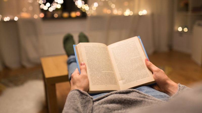 A person reading a book while relaxing indoors, with warm lights in the background during New Year's week. Find financial reads for wealth managers and private bankers.