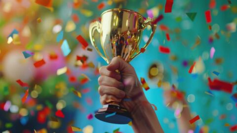 Eine Hand, die eine goldene Trophäe hält, umgeben von buntem Konfetti, symbolisiert Leistung und Exzellenz bei den Private Banking Wealth Awards 2025.