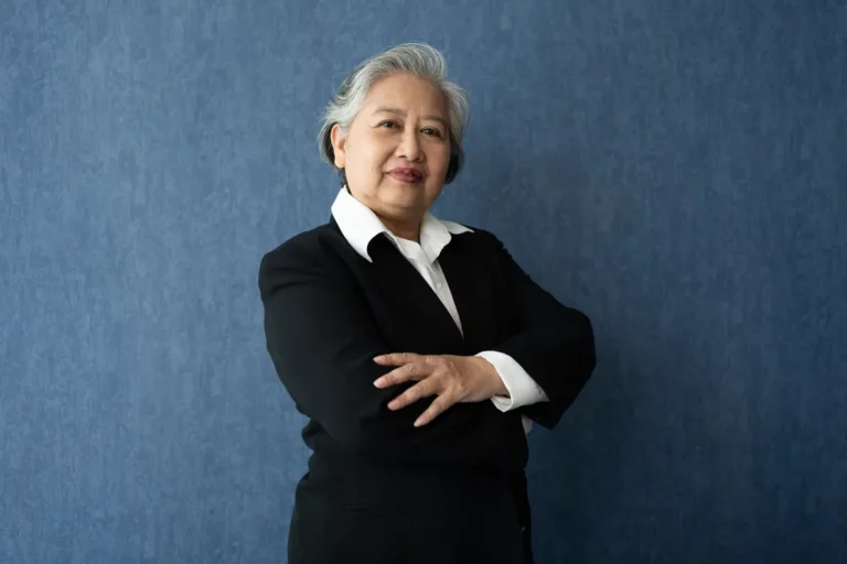 A confident senior financial advisor in a black suit stands with arms crossed against a blue background, symbolizing industry experience.