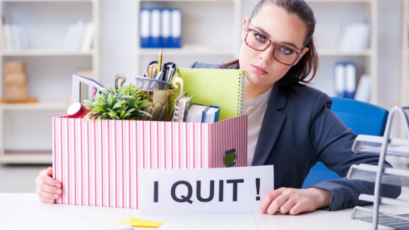 Frau in einem Büro, die eine Kiste mit persönlichen Gegenständen und ein Schild mit der Aufschrift “Ich kündige” in der Hand hält, was die Kündigung eines Arbeitsplatzes symbolisiert.
