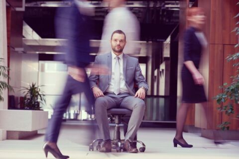 Privater Banker, im stillen Raum sitzend, während Kollegen an ihm vorbeieilen, was die persönliche Beziehung zu Ultra-High-Net-Worth-Kunden inmitten des institutionellen Wandels symbolisiert.