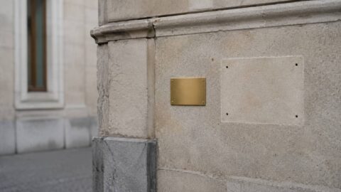 Stone bank facade where a large brass sign has been removed, with a small new blank plate beside it — what Swiss bank mergers leave behind