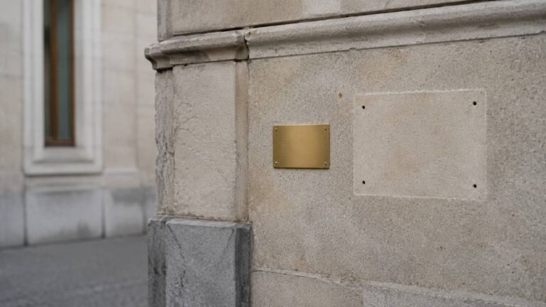 Stone bank facade where a large brass sign has been removed, with a small new blank plate beside it — what Swiss bank mergers leave behind